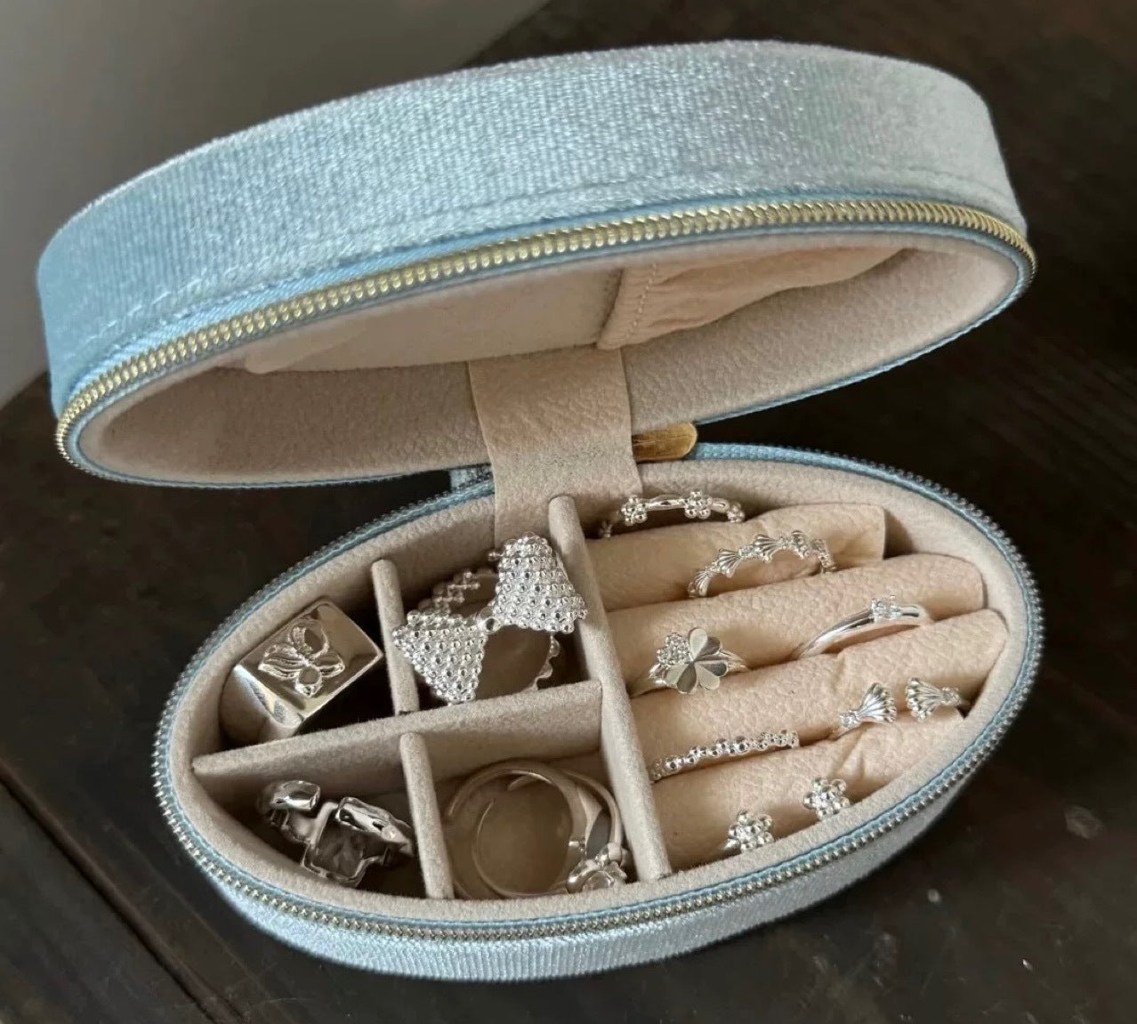 An open oval jewelry organizer case with a glittery light blue exterior and beige interior, holding multiple silver rings and jewelry pieces on padded compartments. The case is photographed on a dark wooden surface.